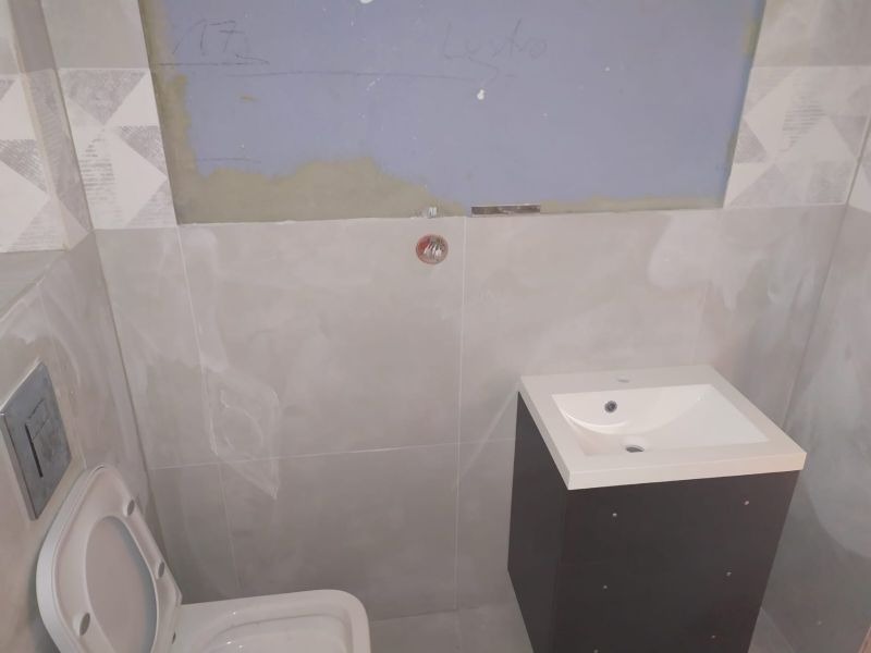 Łazienka w trakcie wykańczania: częściowo położone szare płytki z dekoracyjnym pasem, zamontowana toaleta z otwartą klapą, szafka z umywalką i ślady po pracach na ścianie.