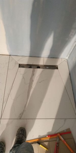 Ułożone białe płytki imitujące marmur z odpływem liniowym w kabinie prysznicowej, widoczne narzędzia glazurnicze i fragment buta wykonawcy.