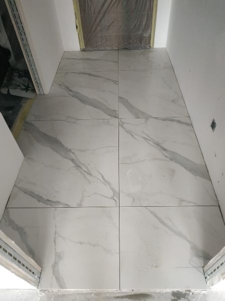 Świeżo ułożone, białe płytki podłogowe z szarym marmurkowym wzorem w wąskim korytarzu podczas prac wykończeniowych, widoczne profile przy ścianach i zabezpieczone drzwi.