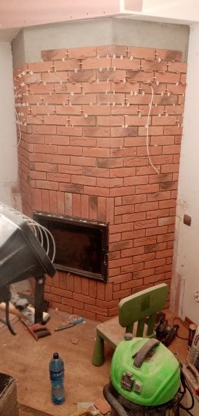 Montaż okładziny ceglanej wokół wkładu kominkowego w rogu pokoju, widoczne krzyżyki dystansowe i fragmenty narzędzi na podłodze.