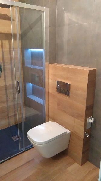 Nowoczesna łazienka z podwieszaną toaletą na drewnianej zabudowie, szklaną kabiną prysznicową z wnękami podświetlonymi LED, wykończona płytkami imitującymi drewno.