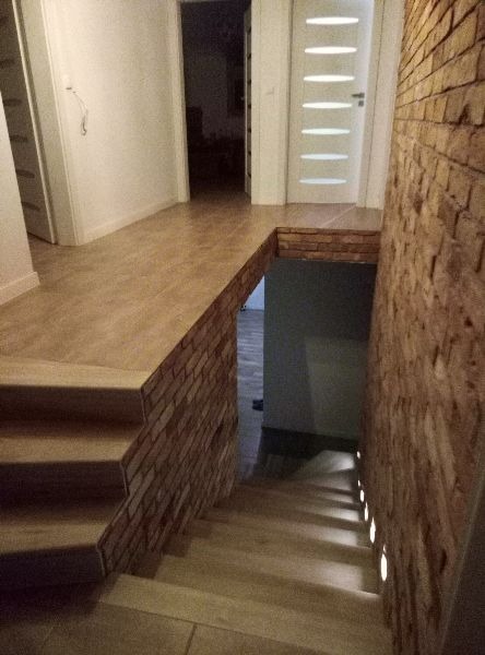 Wykończone wnętrze domu z drewnianą podłogą, schodami z podświetleniem LED i ścianą z cegły dekoracyjnej, widok na białe drzwi z przeszkleniami i otwarte przejście do dalszej części domu.