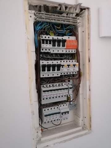 Otwarta skrzynka elektryczna w ścianie z widocznymi przewodami i bezpiecznikami, w trakcie prac instalacyjnych.