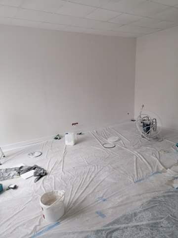 Pokój przygotowany do malowania: białe ściany, podłoga zabezpieczona folią malarską, widoczne pędzle, wałki i wiadro z farbą w rogu pomieszczenia oraz agregat malarski.