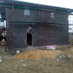 Niedokończony dom z czarną elewacją z izolacji, otoczony rusztowaniami, z widocznym stosem piasku i gruzu na trawiastym terenie.