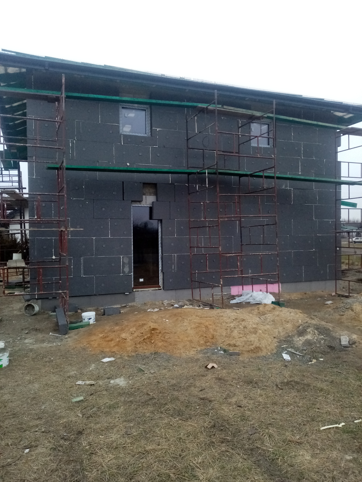 Niedokończony dom z czarną elewacją z izolacji, otoczony rusztowaniami, z widocznym stosem piasku i gruzu na trawiastym terenie.