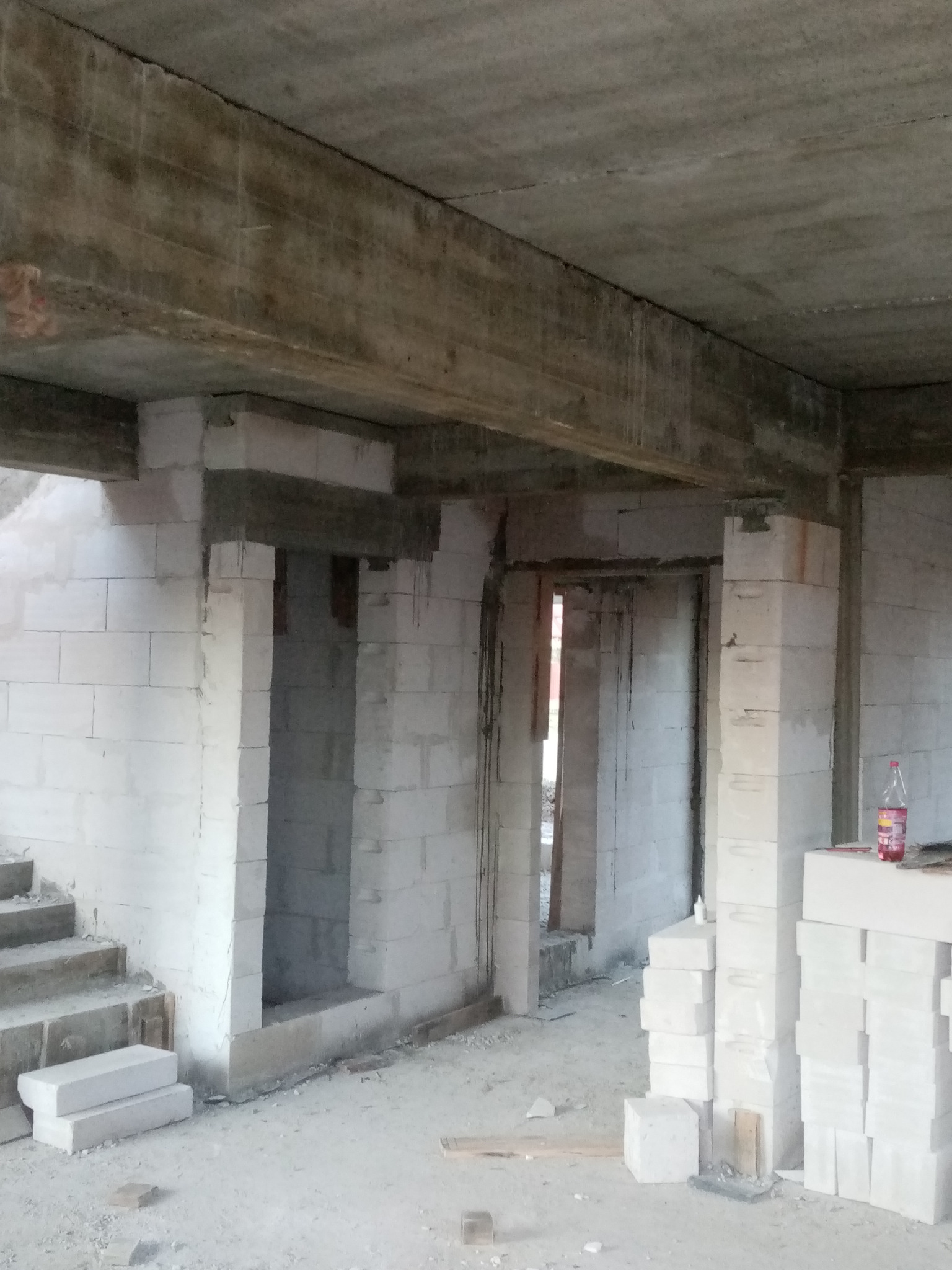 Wnętrze surowego domu w trakcie budowy z betonu komórkowego, widoczne surowe betonowe stropy, schody, otwory drzwiowe oraz butelka z napojem na murze.