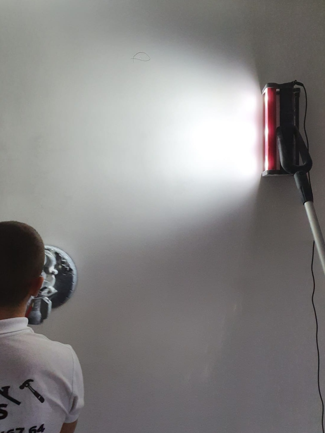 Pracownik szlifuje ścianę za pomocą szlifierki z oświetleniem LED, widoczny pył unoszący się w powietrzu pod wpływem światła.