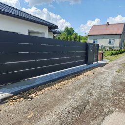 AL-DO Piotr Haśkiewicz - Nowoczesna, przesuwna brama w kolorze antracytu z poziomymi panelami, zamontowana na posesji z kostką brukową i kamieniami. Widoczny automat do bramy.