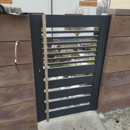 AL-DO Piotr Haśkiewicz - Nowoczesna, grafitowa furtka z poziomymi szczeblami, wkomponowana w ogrodzenie z brązowych, betonowych bloczków. Widoczne okablowanie sugeruje etap prac wykończeniowych.