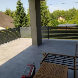 Nowoczesny balkon z szarą, poziomą balustradą z metalowych profili, szarą podłogą z płytek i widokiem na zieleń drzew; w rogu widoczne narzędzia i skrzynka narzędziowa.
