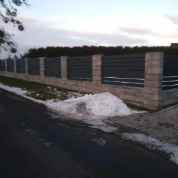 Nowoczesne ogrodzenie panelowe z betonowymi słupkami i poziomymi, grafitowymi przęsłami, widoczne z ulicy w zimowej scenerii z resztkami śniegu.