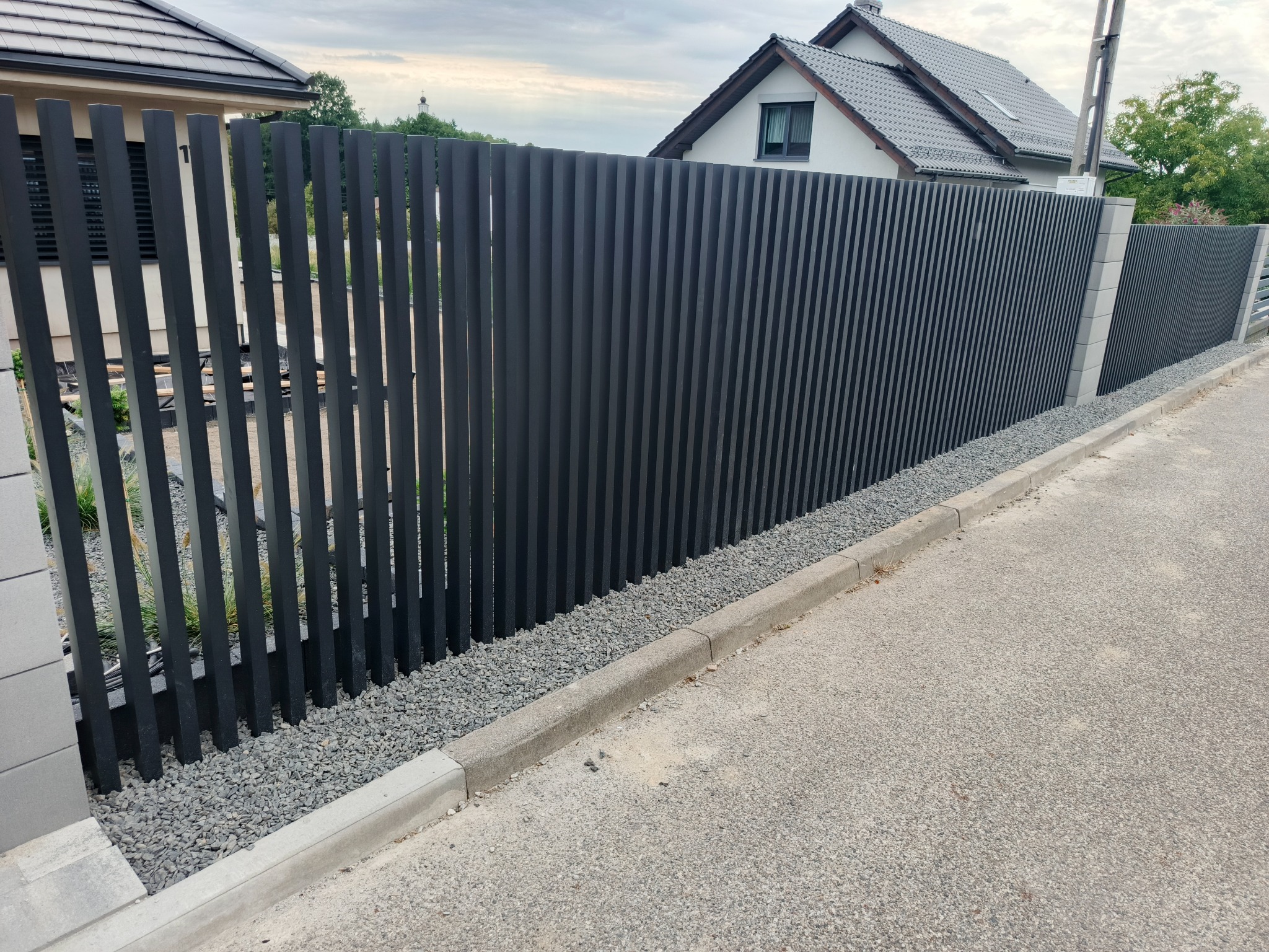 Nowoczesne, pionowe ogrodzenie panelowe w kolorze antracytowym, oddzielające posesję od drogi. Widoczne szare kamienie dekoracyjne u podstawy i fragment domu w tle.