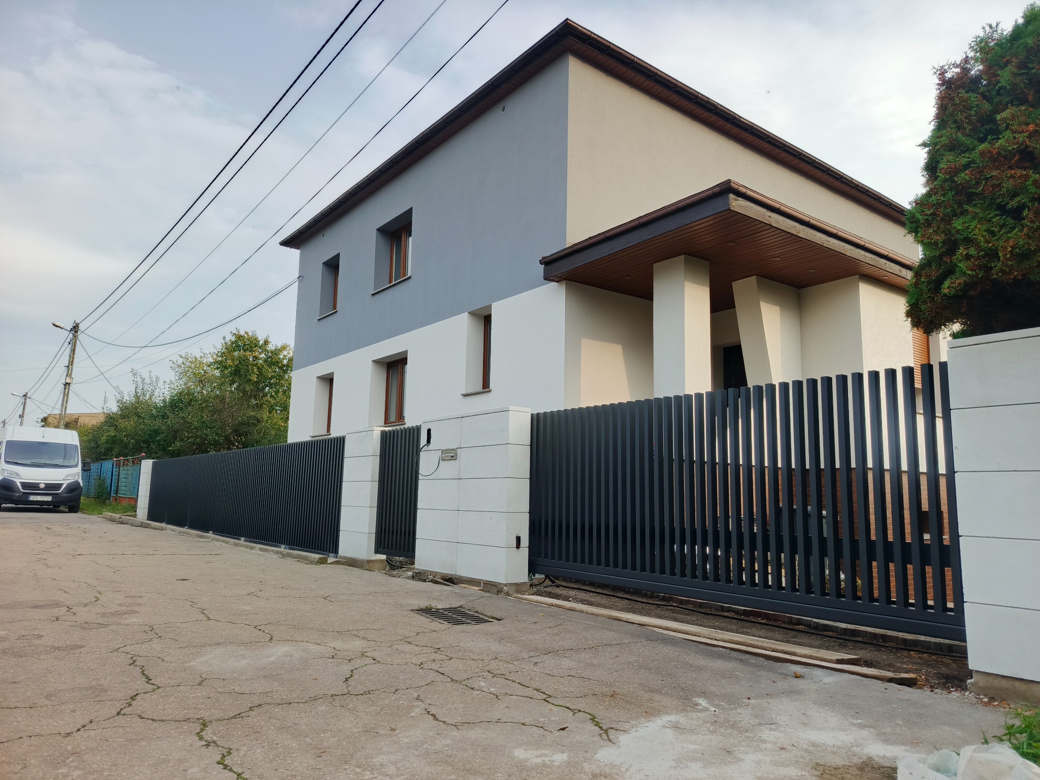 Nowoczesna, przesuwna brama wjazdowa z pionowych, grafitowych profili, dopasowana do ogrodzenia i architektury domu. Widoczny automat do bramy.