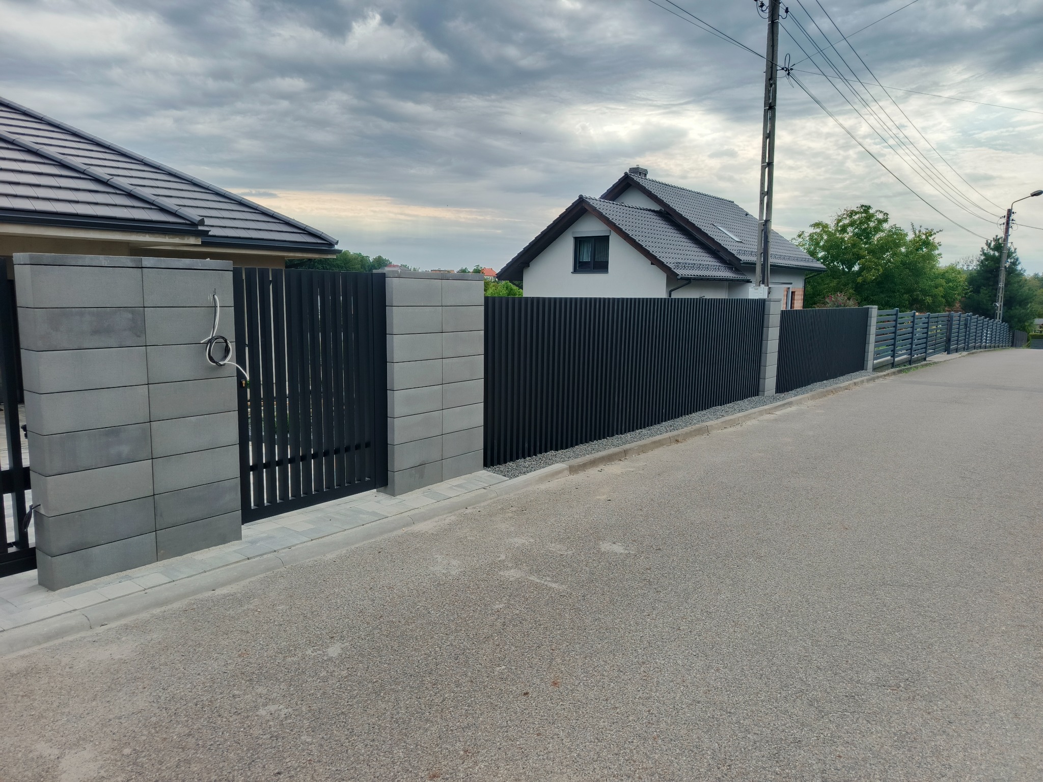 Nowoczesne ogrodzenie panelowe w kolorze antracytowym z betonowymi słupkami, widoczne na tle domów jednorodzinnych i pochmurnego nieba. Minimalistyczny design.