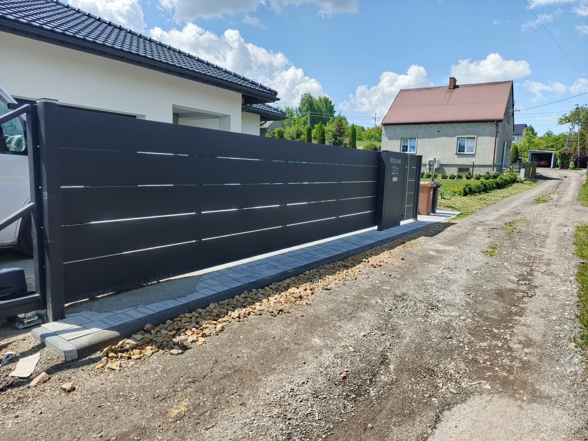 Nowoczesna, przesuwna brama w kolorze antracytu z poziomymi panelami, zamontowana na posesji z kostką brukową i kamieniami. Widoczny automat do bramy.