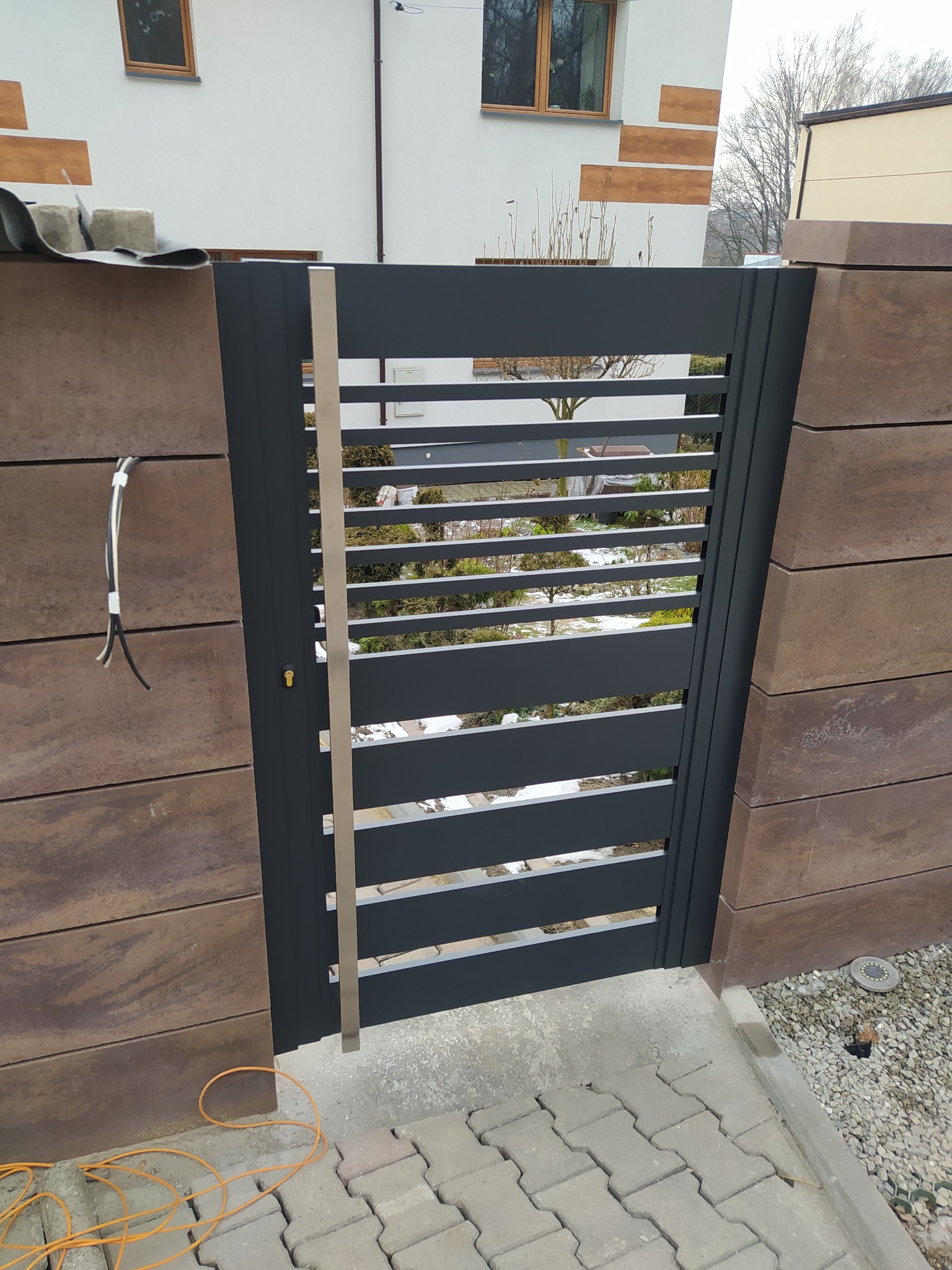 Nowoczesna, grafitowa furtka z poziomymi szczeblami, wkomponowana w ogrodzenie z brązowych, betonowych bloczków. Widoczne okablowanie sugeruje etap prac wykończeniowych.