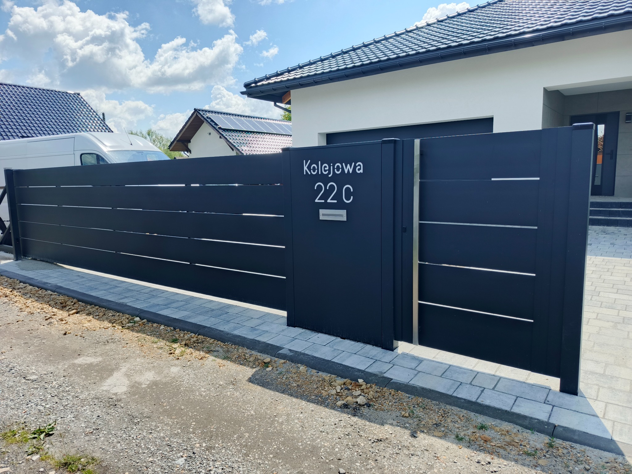 Nowoczesna, antracytowa brama wjazdowa z numerem posesji 'Kolejowa 22 C' na tle domu z ciemną dachówką i błękitnego nieba z chmurami. Minimalistyczny design.