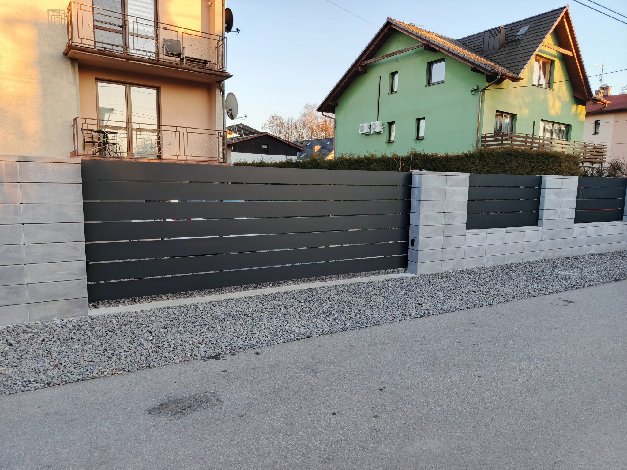 Nowoczesne ogrodzenie posesji z poziomych, grafitowych paneli, osadzone na szarych, betonowych słupkach. Tło: domy w zabudowie jednorodzinnej i szara droga.