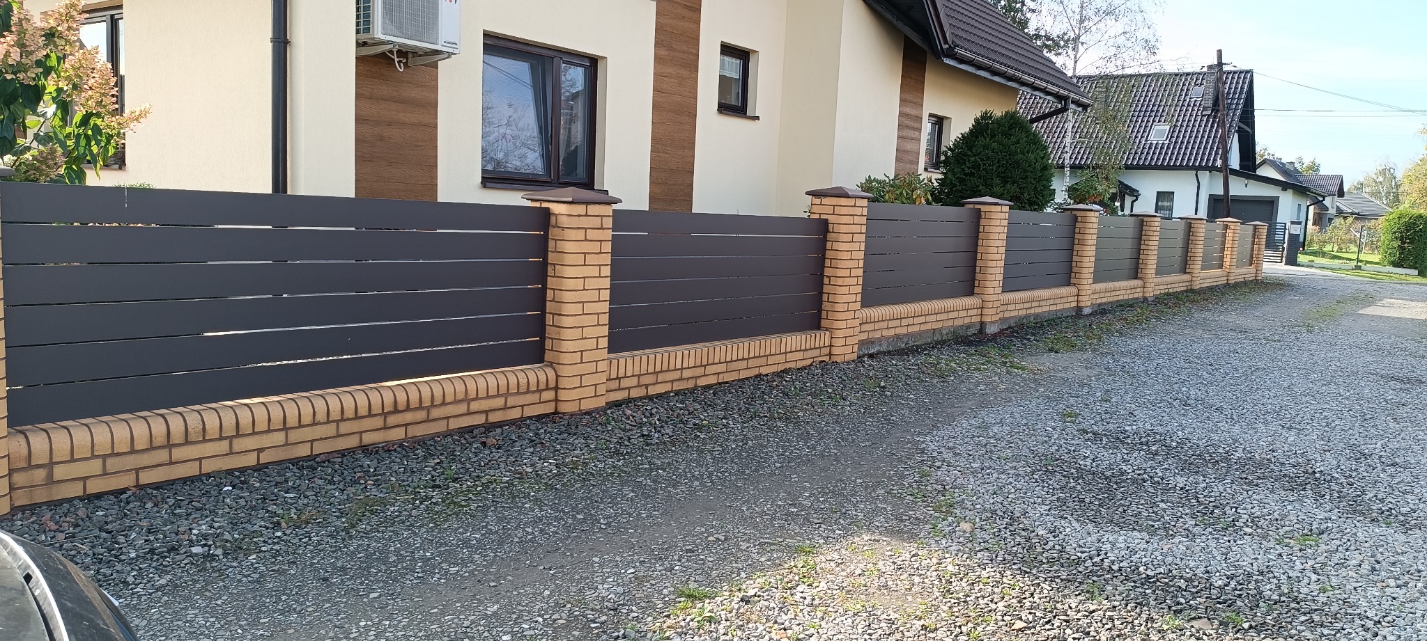 Nowoczesne ogrodzenie posesji z poziomych, szarych paneli, wsparte na ceglanych słupkach i podmurówce, na tle domu z klimatyzatorem i żwirowego podjazdu.