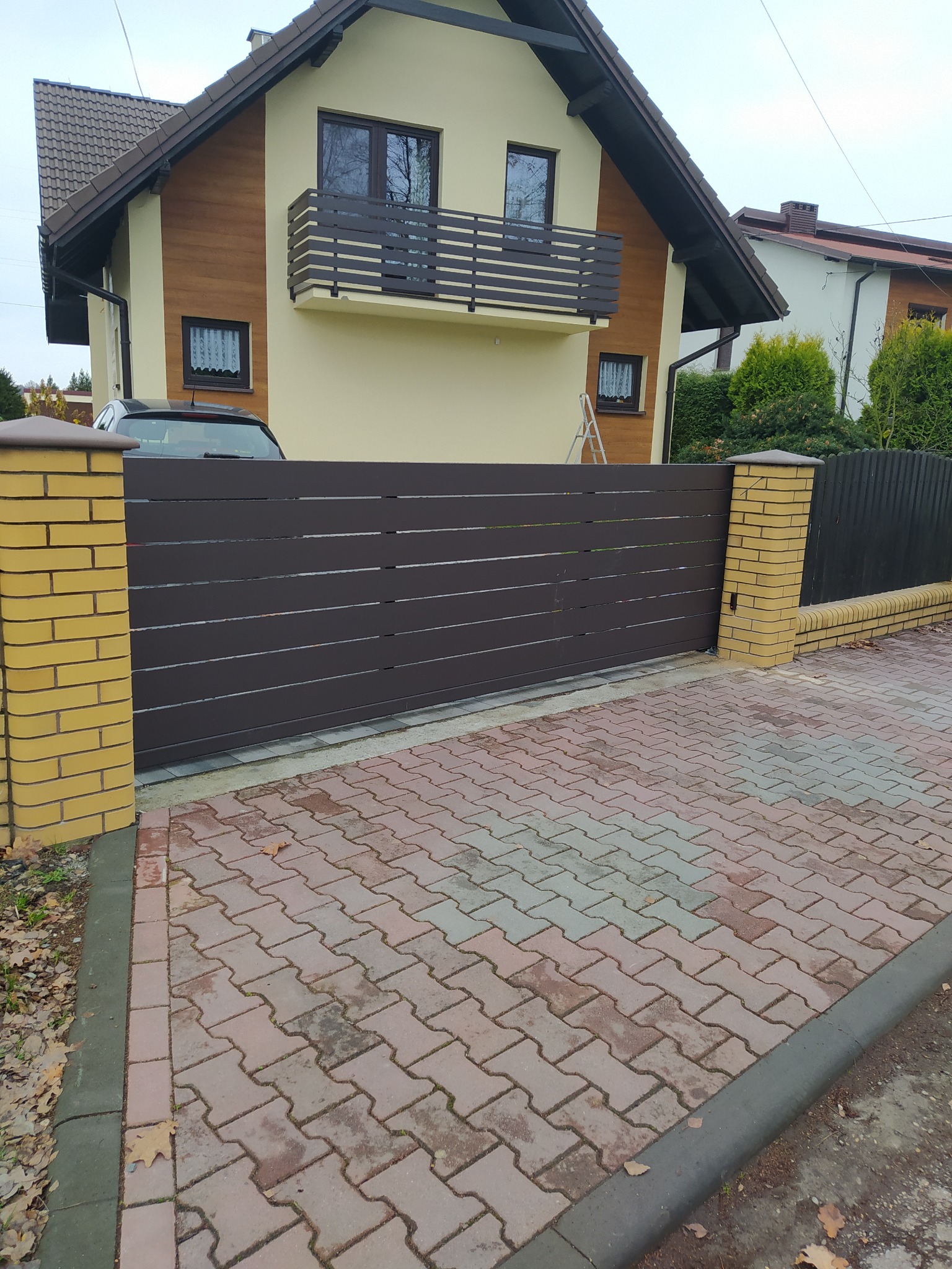 Nowoczesna, pozioma brama przesuwna w kolorze brązowym, z ceglanymi słupkami, na tle domu z balkonem i kostki brukowej. Widok z zewnątrz.