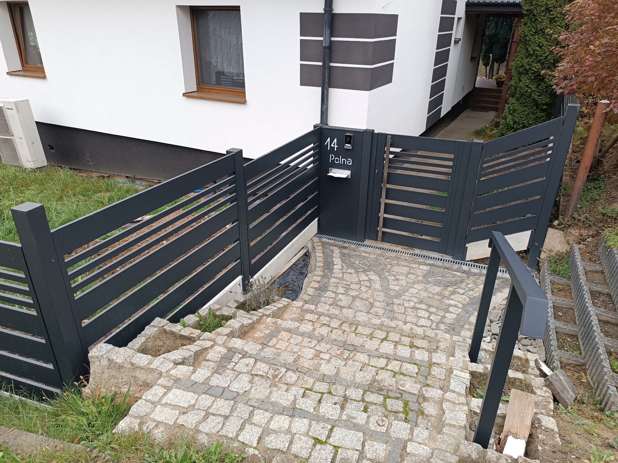 Nowoczesne, poziome ogrodzenie panelowe w kolorze antracytowym z furtką i numerem domu, prowadzące do wejścia po schodach z kostki brukowej.