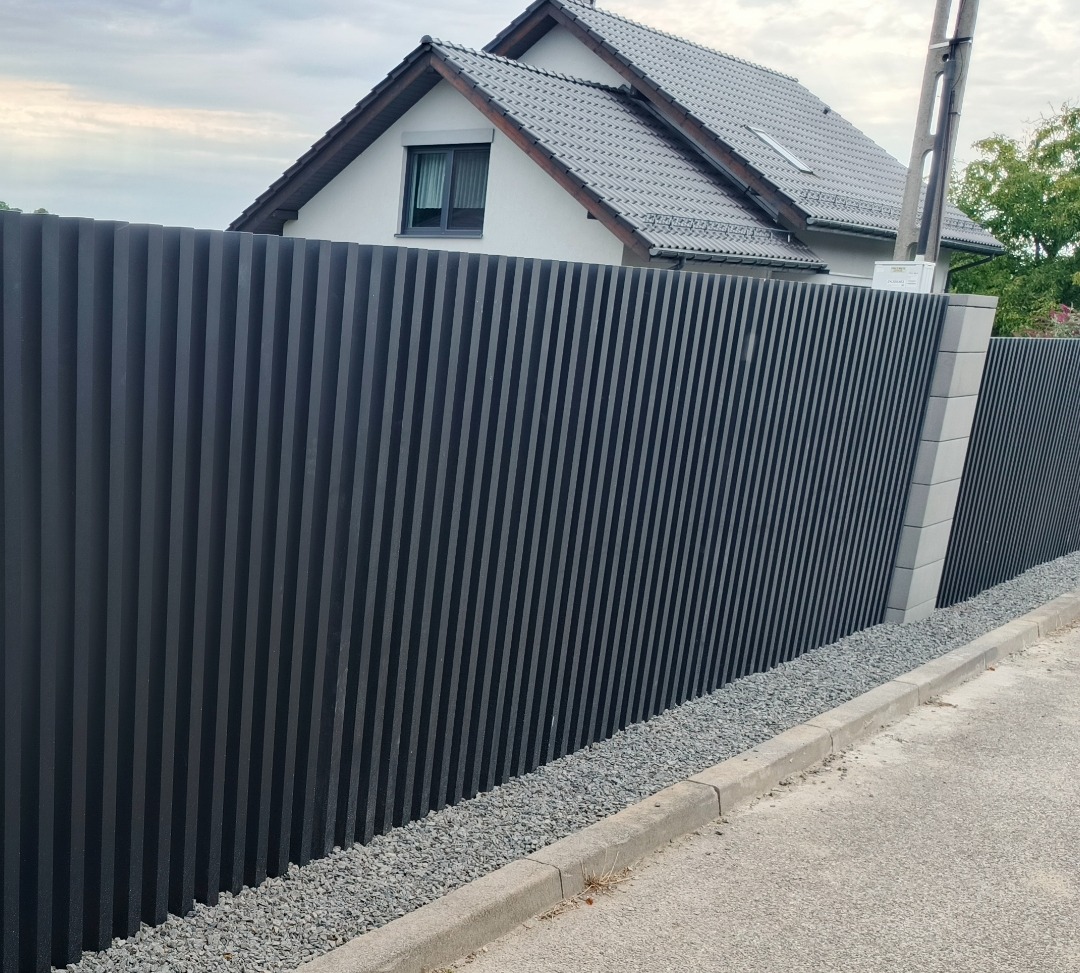 Nowoczesne, pionowe ogrodzenie metalowe w kolorze grafitowym, z betonowymi słupkami, na tle domu z szarą dachówką i pochmurnego nieba. Minimalistyczny design.