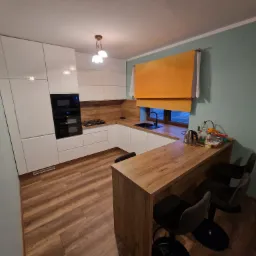 Nowoczesna kuchnia z białymi, błyszczącymi frontami szafek, drewnianym blatem i barkiem z hokerami, oświetlona lampą z trzema kloszami.