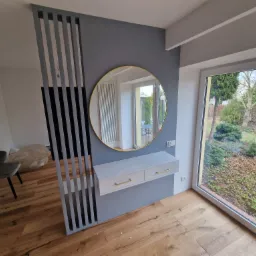 Nowoczesna aranżacja przedpokoju z drewnianą podłogą, szarą ścianą, okrągłym lustrem w złotej ramie, białą konsolą z szufladami i ażurową ścianką działową.