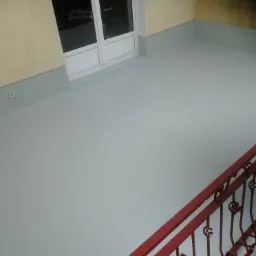 Szary balkon z pomalowaną podłogą, widoczne czerwone, metalowe barierki i białe drzwi balkonowe.