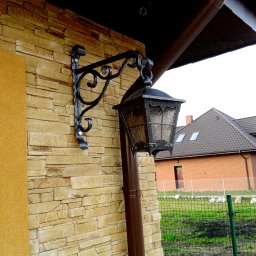 DOMIX JanPrzystański - Kuta, ozdobna lampa elewacyjna z metalową konstrukcją, zamontowana na ścianie z kamienia dekoracyjnego, widoczny fragment rynny i domu w tle.