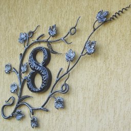 DOMIX JanPrzystański - Kuta, metalowa numeracja domu w kształcie cyfry 8, otoczona stylizowanymi pędami winorośli z liśćmi, zamontowana na teksturowanej, beżowej elewacji budynku.