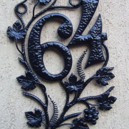 DOMIX JanPrzystański - Kuta, czarna tabliczka z numerem domu '64' opleciona motywem winorośli z liśćmi i gronami, zamontowana na teksturowanej, beżowej ścianie.