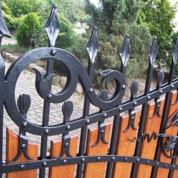 DOMIX JanPrzystański - Fragment ogrodzenia z drewnianymi panelami i kutymi, czarnymi zdobieniami w kształcie liści winogron i spiral, z zielenią w tle.