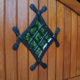 Fragment drewnianej bramy z ozdobnym, kutym okienkiem w kształcie rombu, przez które widać zieleń.