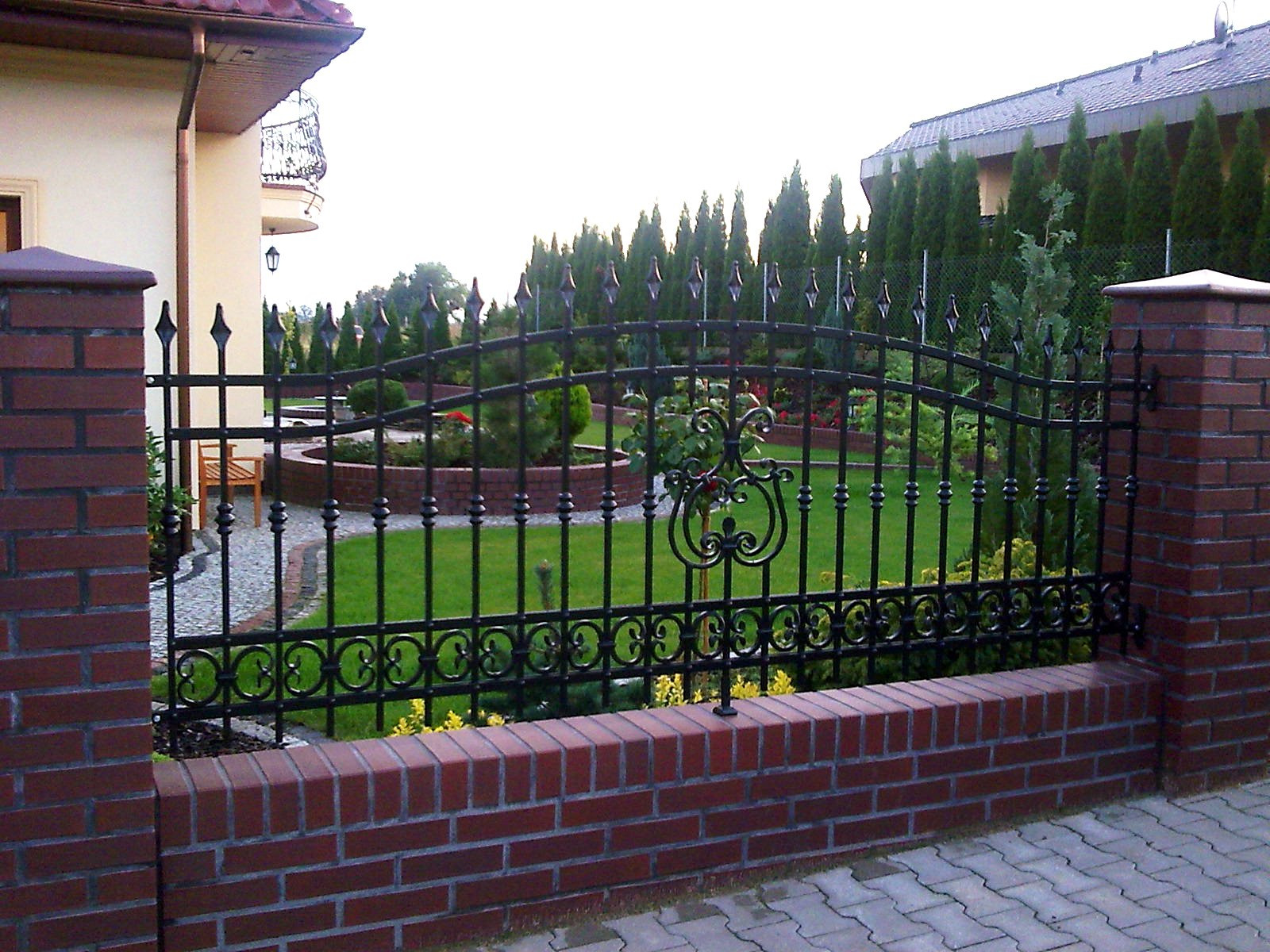 Eleganckie, czarne ogrodzenie z kutego żelaza, osadzone na niskim murku z czerwonej cegły, z widokiem na zadbany ogród z tujami i drewnianą ławką w tle.