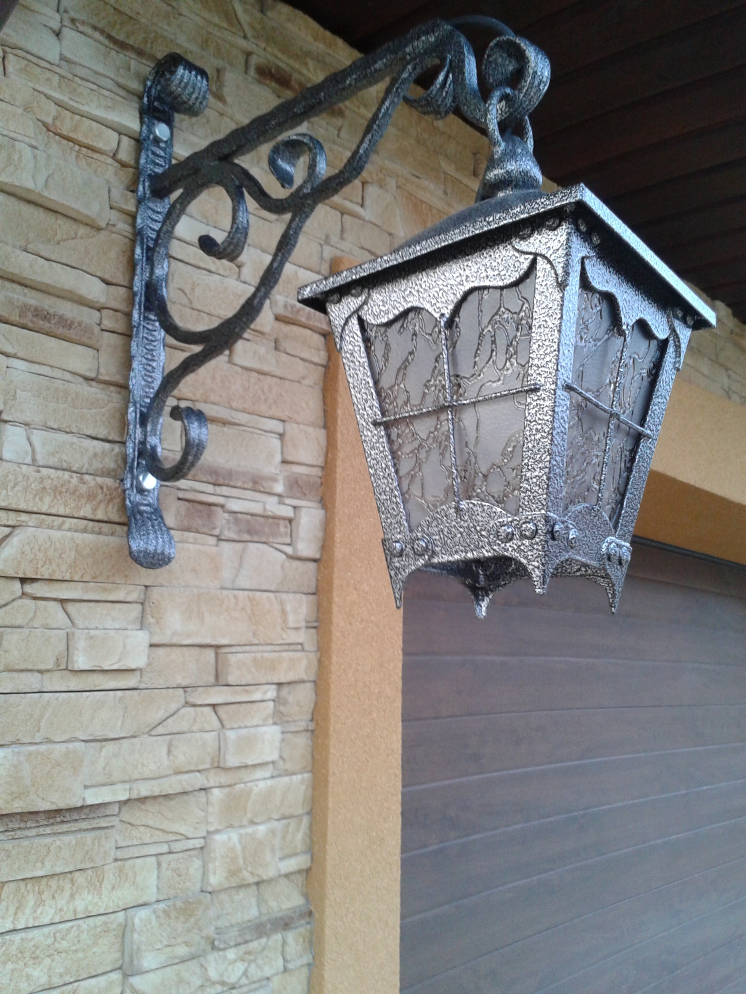 Kuta, ozdobna lampa zewnętrzna z metalu, zamocowana na elewacji z kamienia dekoracyjnego, z widoczną bramą garażową w tle.