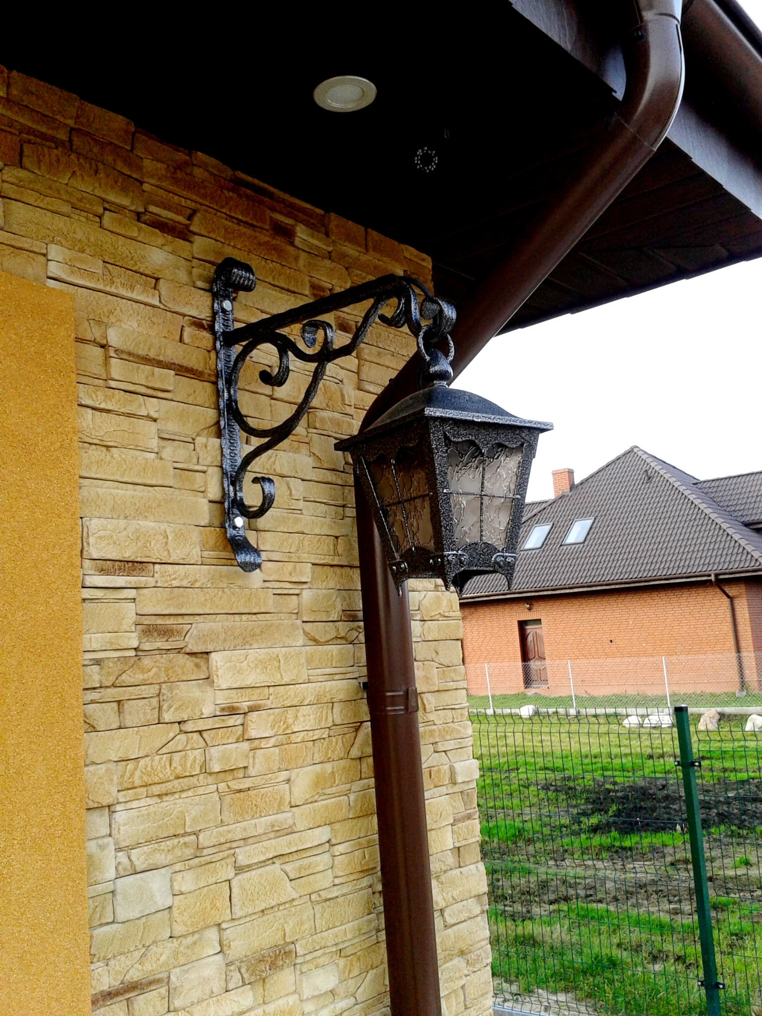 Kuta, ozdobna lampa elewacyjna z metalową konstrukcją, zamontowana na ścianie z kamienia dekoracyjnego, widoczny fragment rynny i domu w tle.