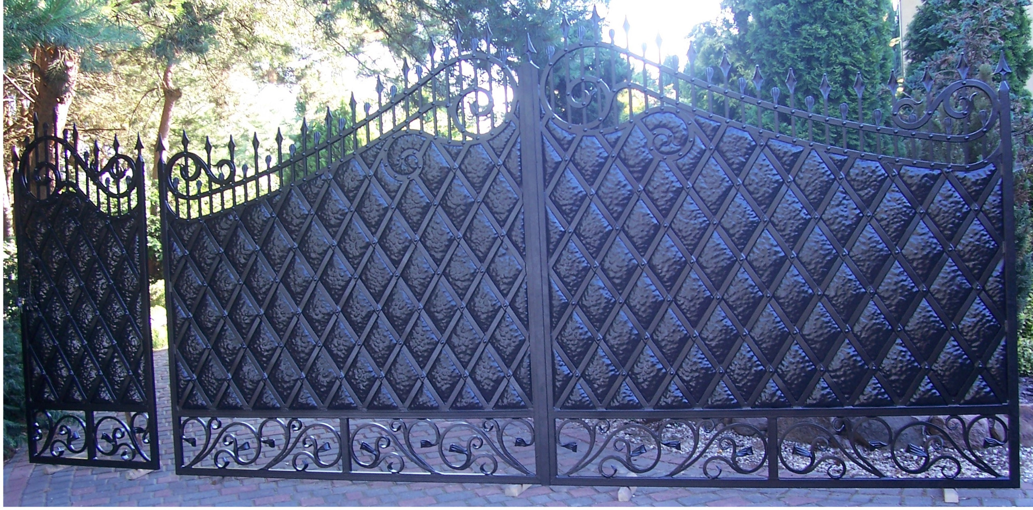 Otwarta dwuskrzydłowa brama z czarnego metalu o dekoracyjnym romboidalnym wzorze i zdobieniach u góry i dołu, z fragmentem ogrodzenia po lewej stronie, na tle zieleni.