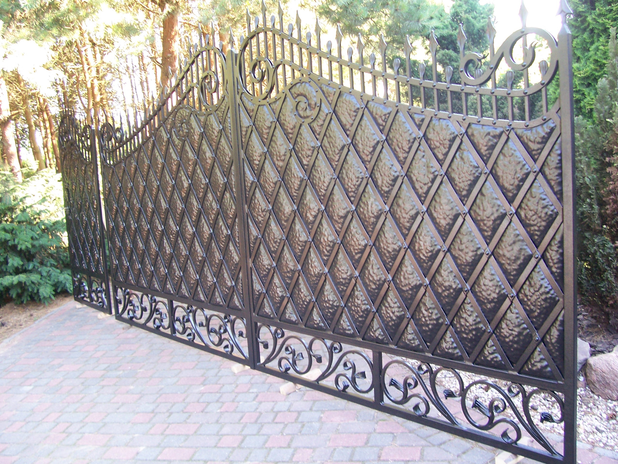 Otwarta, dwuskrzydłowa brama wjazdowa kuta z romboidalnym wypełnieniem i ozdobnymi elementami roślinnymi na tle zieleni i kostki brukowej.