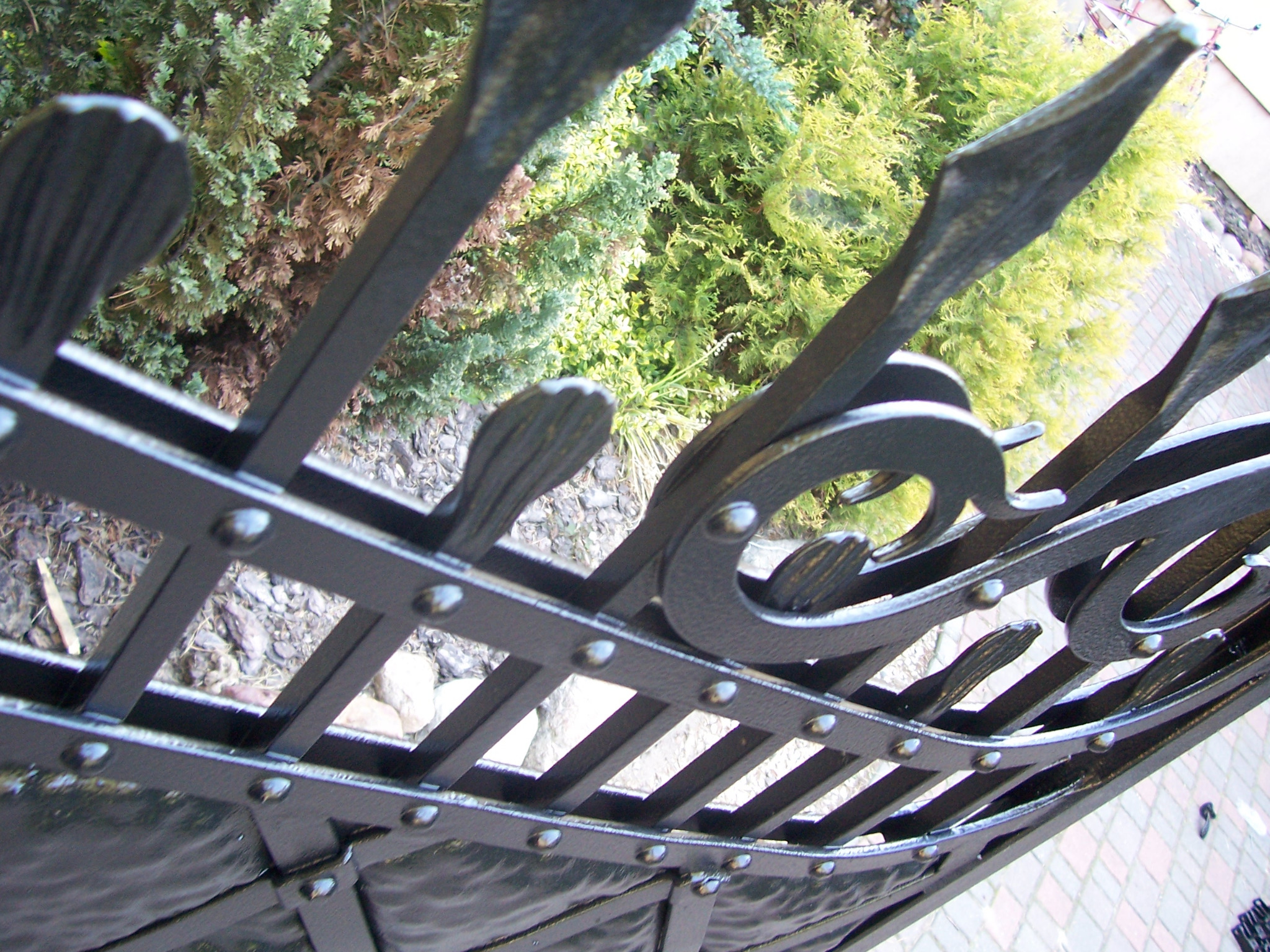 Fragment czarnego ogrodzenia z metalu z dekoracyjnymi elementami i nitami, widzianego z bliska pod kątem, z zielenią w tle.