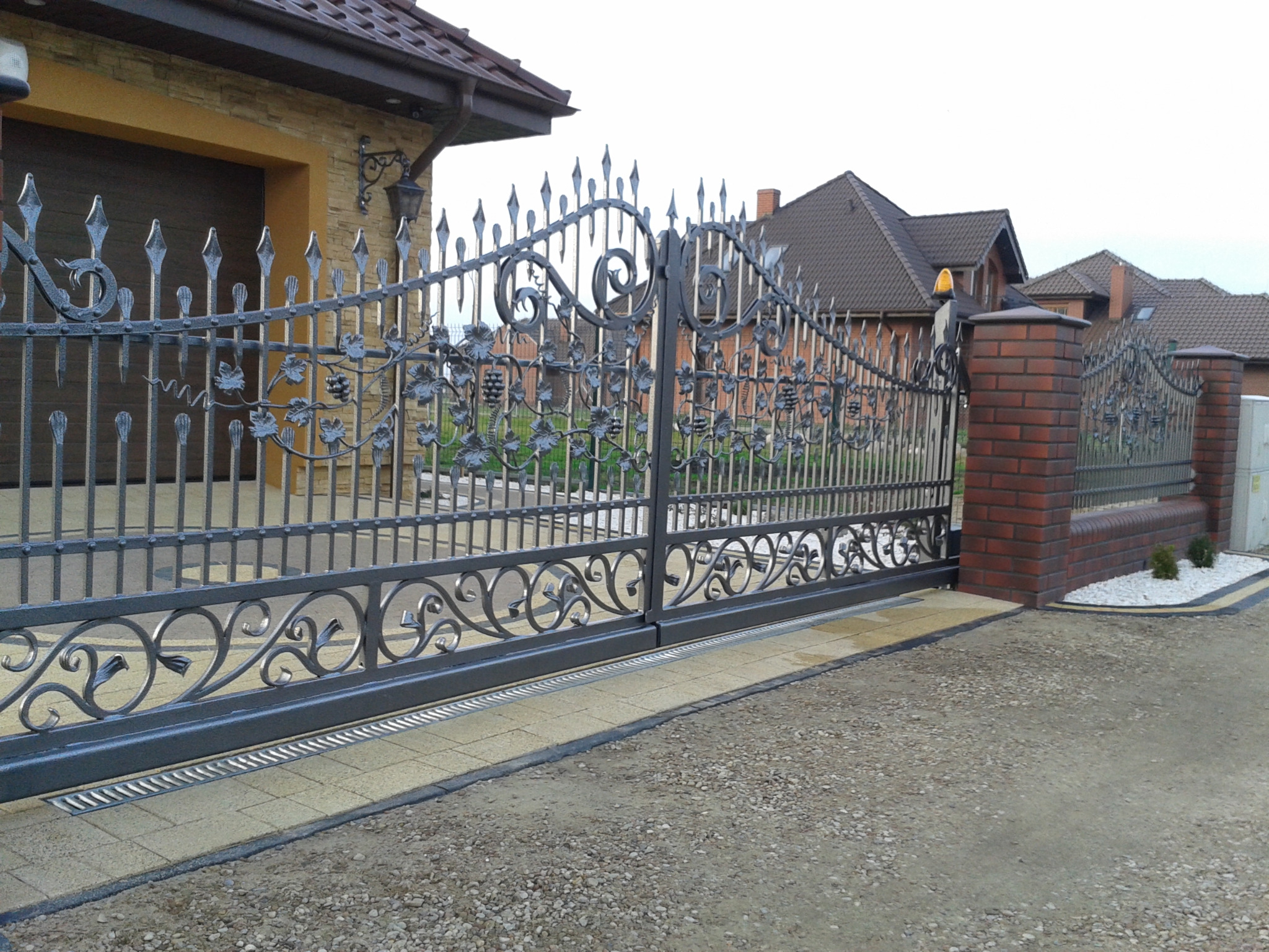 Automatyczna brama przesuwna z metaloplastyki, wzór z motywem winogron, widoczna studzienka odpływowa liniowa, w tle dom jednorodzinny z garażem.