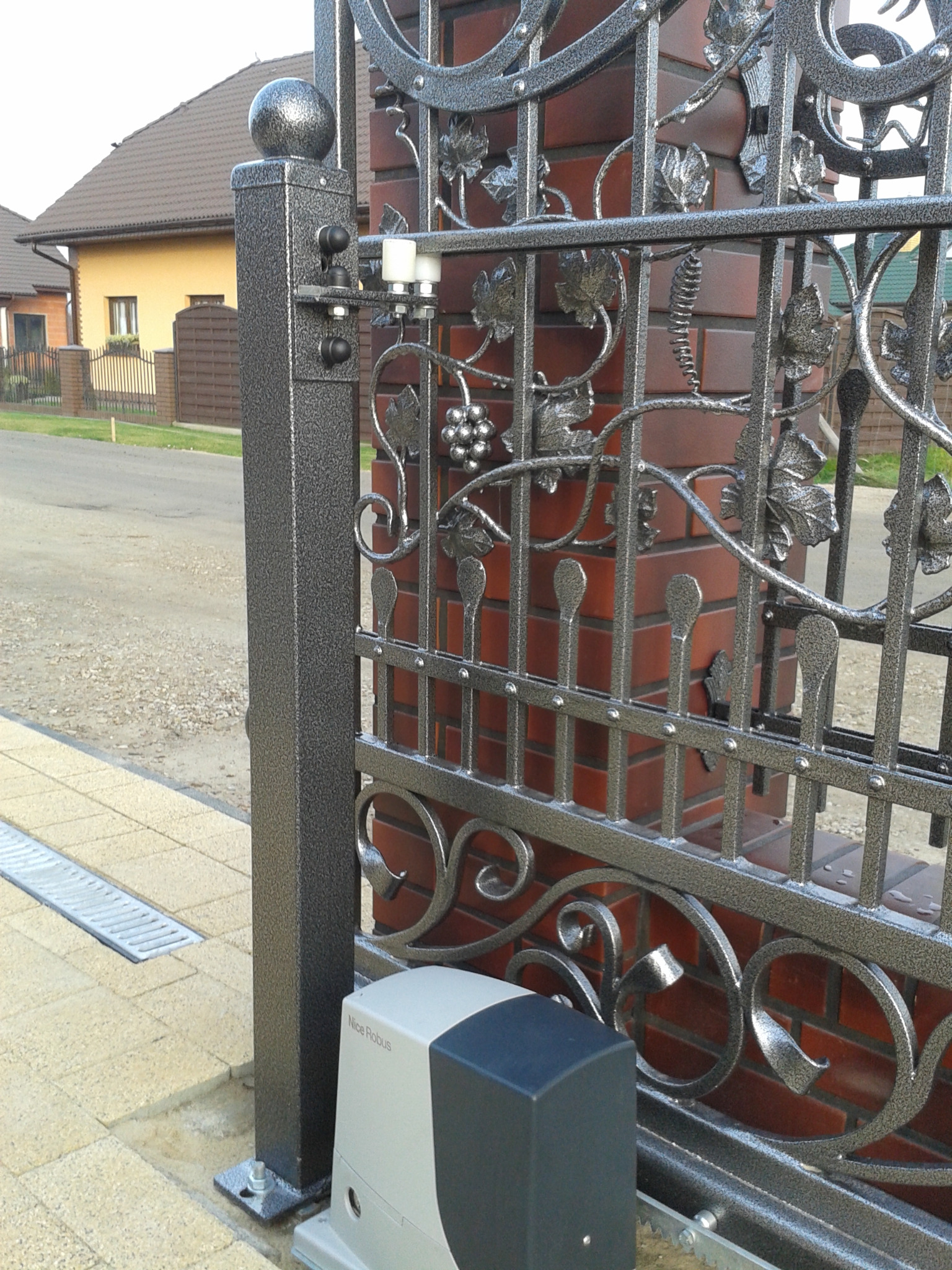 Fragment automatycznej bramy wjazdowej z metaloplastyki, motyw winogron i liści, zamontowany napęd Nice Robus, słupek z kulą na szczycie, ceglany filar, kostka brukowa.