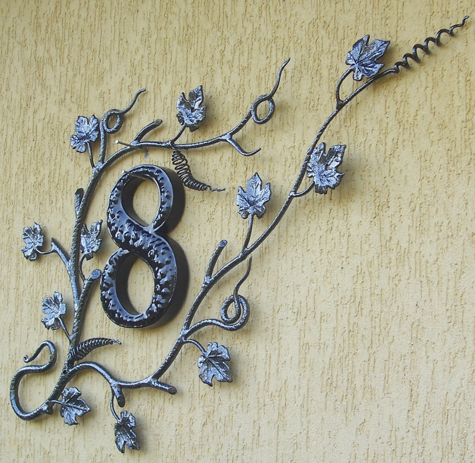 Kuta, metalowa numeracja domu w kształcie cyfry 8, otoczona stylizowanymi pędami winorośli z liśćmi, zamontowana na teksturowanej, beżowej elewacji budynku.