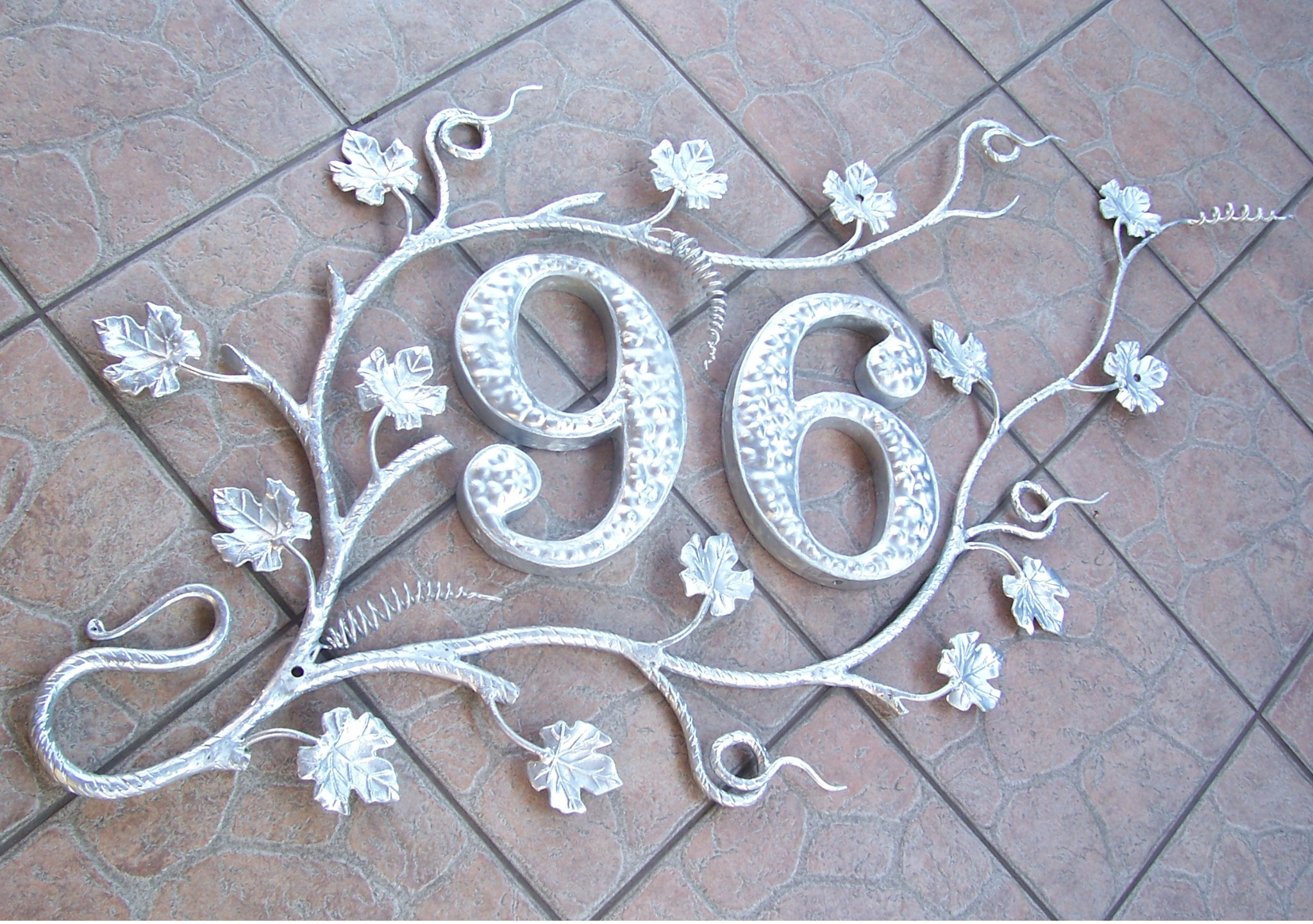 Metalowy numer domu '96' w stylu roślinnym z liśćmi klonu, umieszczony na brązowych płytkach chodnikowych.