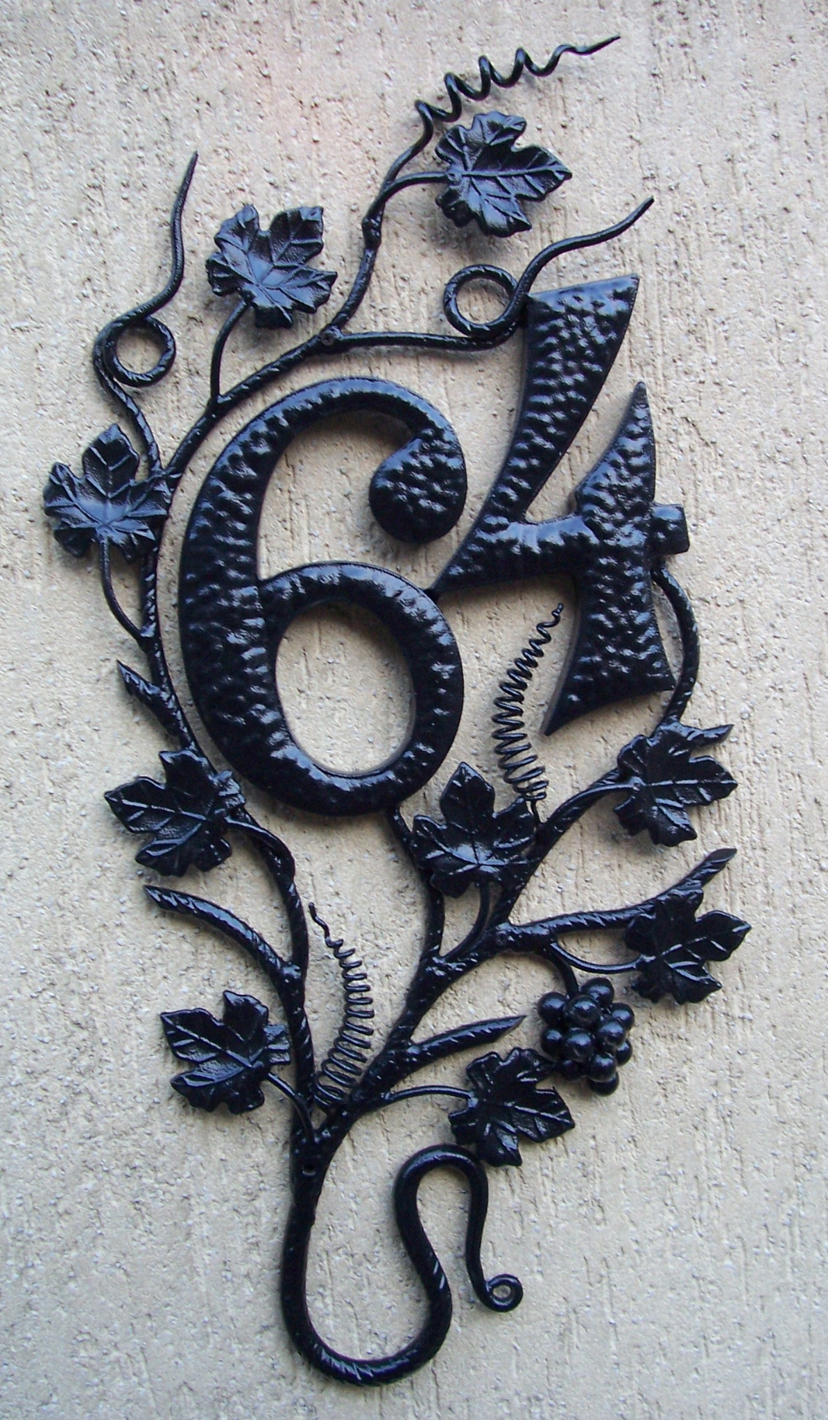 Kuta, czarna tabliczka z numerem domu '64' opleciona motywem winorośli z liśćmi i gronami, zamontowana na teksturowanej, beżowej ścianie.