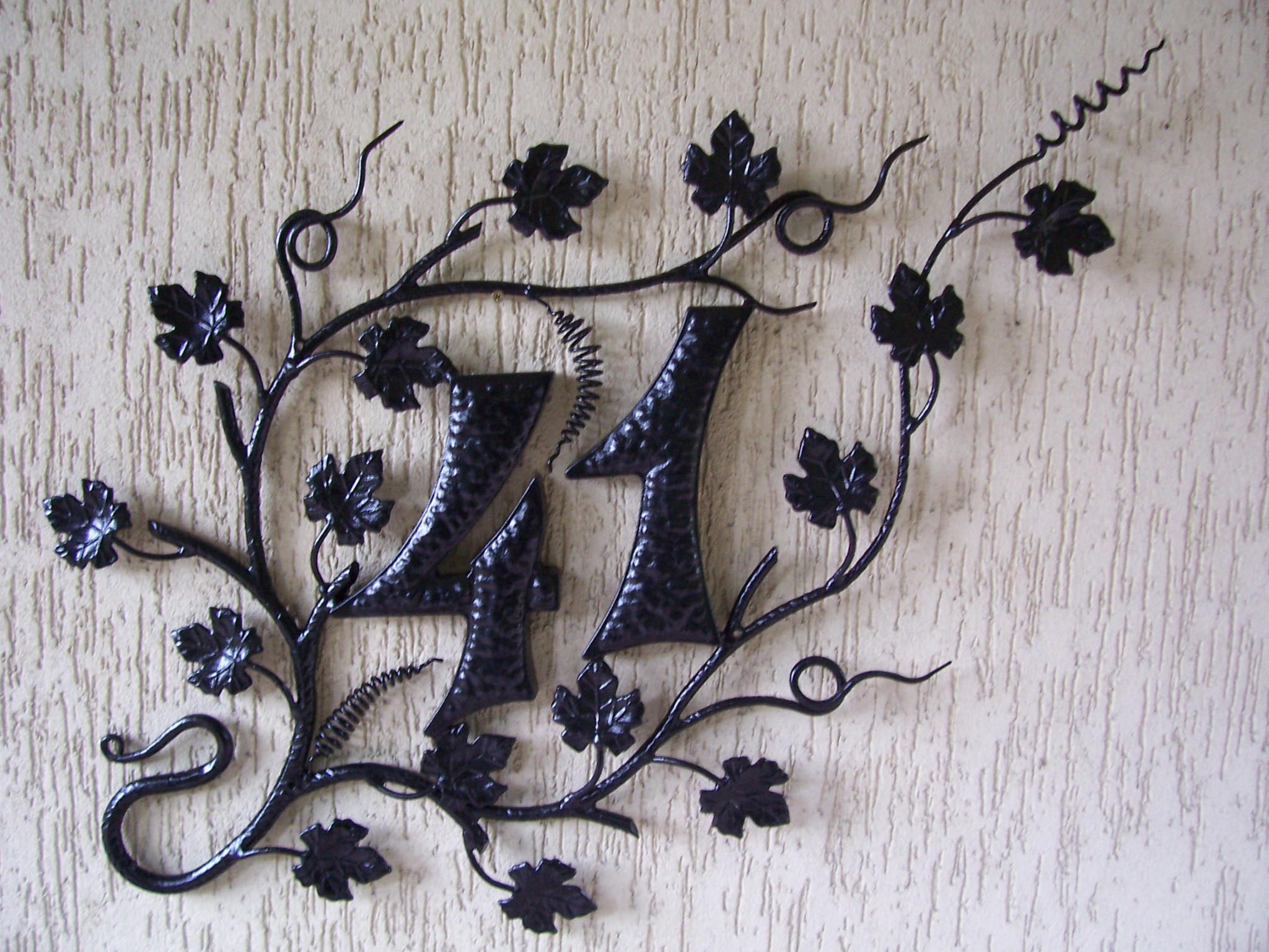 Numer domu '44' wykonany z metaloplastyki, stylizowany na pnącze winorośli z liśćmi, zamontowany na teksturowanej ścianie.