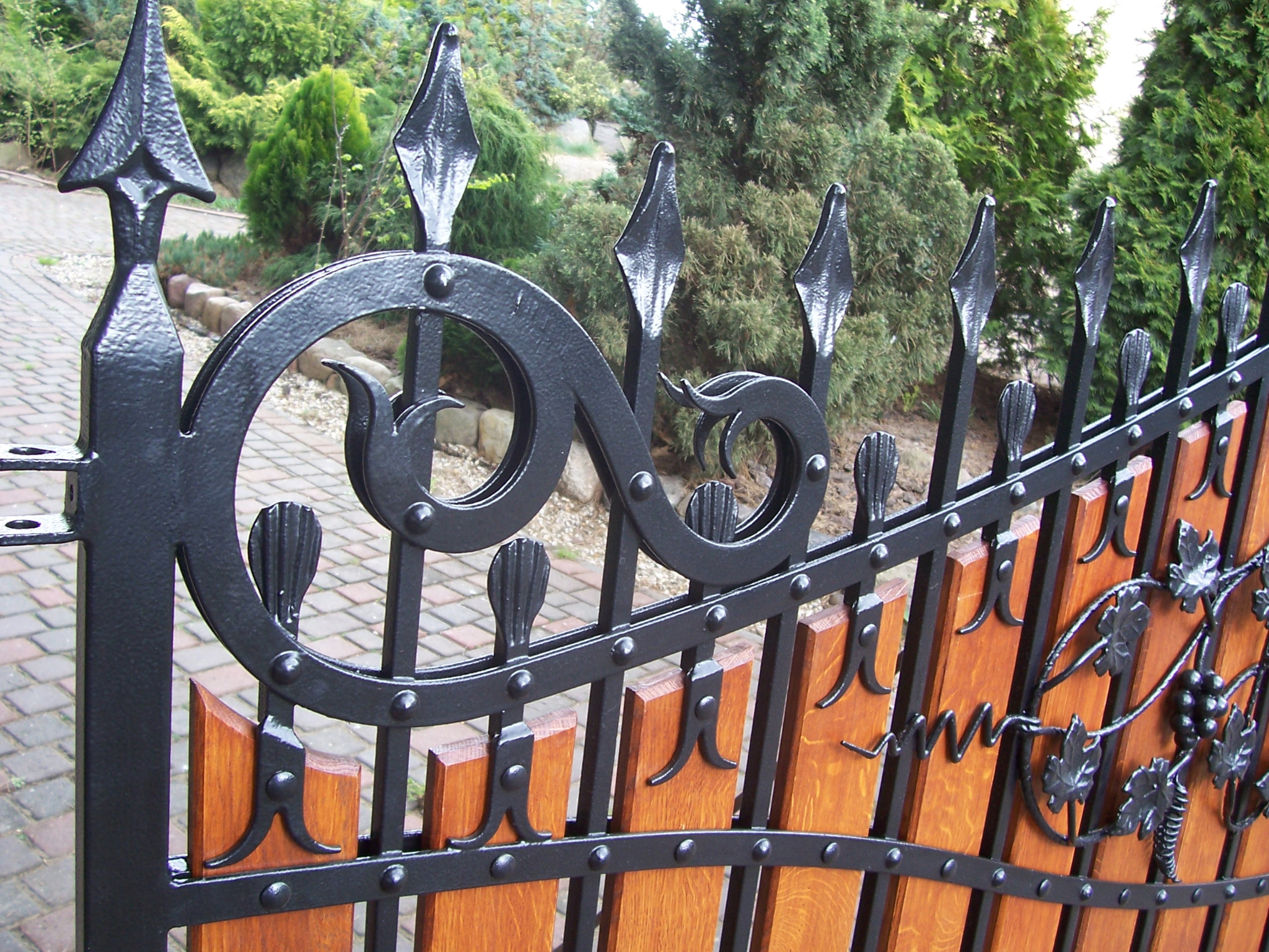 Fragment ogrodzenia z drewnianymi panelami i kutymi, czarnymi zdobieniami w kształcie liści winogron i spiral, z zielenią w tle.