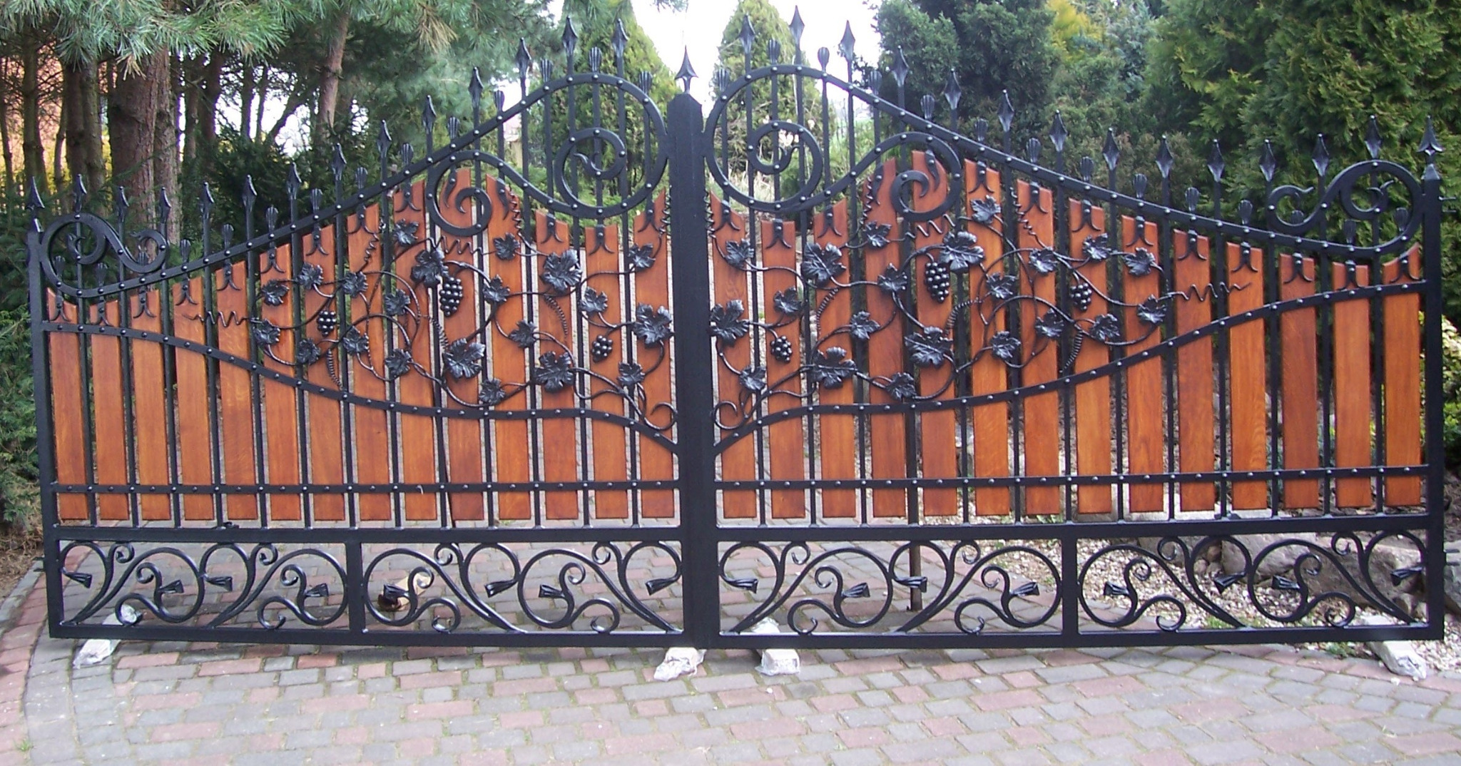 Dwuskrzydłowa brama z drewnianym wypełnieniem i kutymi zdobieniami z motywem winogron, widoczna z zewnątrz na tle zieleni.