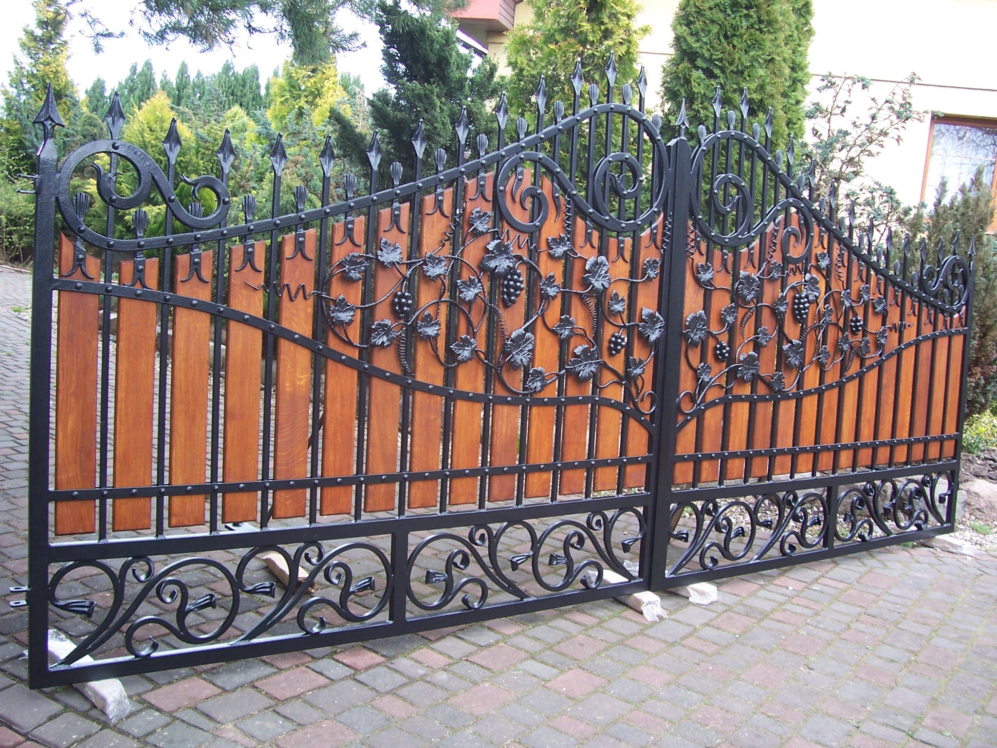 Dwuskrzydłowa brama wjazdowa z czarnego metalu z dekoracyjnymi elementami roślinnymi (liście winogron i winogrona) i drewnianym wypełnieniem, na posesji wyłożonej kostką brukową.